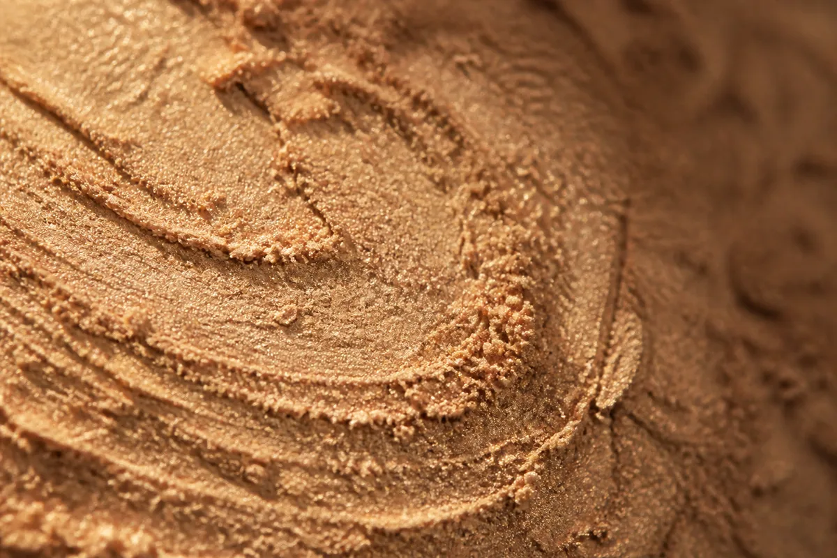 Gros plan sur une texture de poudre minérale beige tamisée par la lumière rasante